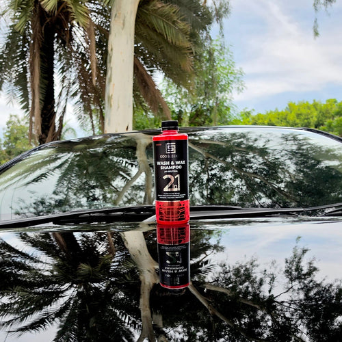 A bottle of 2 in 1 Wash & Shine car wash product standing on the windshield of a black car, with palm trees and a cloudy sky reflected in the wet surface.